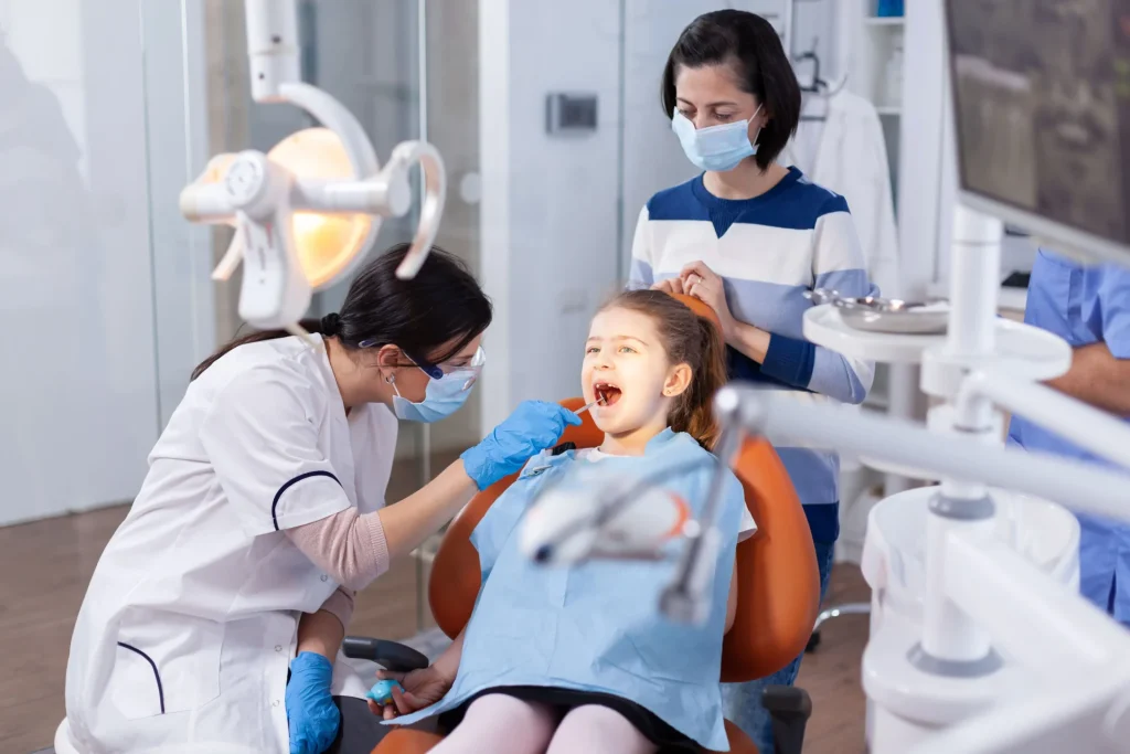 Criança sentada em cadeira de Dentista sendo examinada com um instrumento que possui espelho por uma Dentista e sendo acompanhada pela sua mãe. 