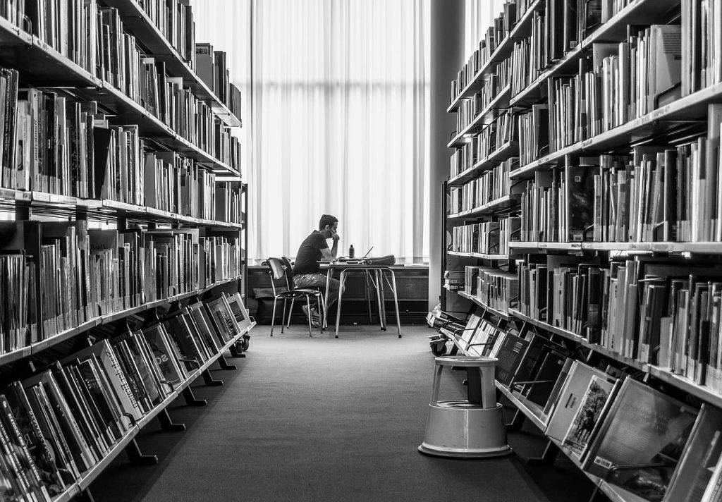 Pessoa sentada, lendo um livro em uma espécie de biblioteca com um corredor que possui muitas prateleiras com livros. 